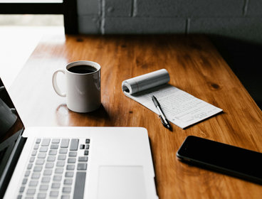 An open laptop, coffee mug containing coffee, notebook, pen and mobile phone on a desk.