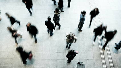 Several individuals walking swiftly on a grey concrete ground whist four individuals stand stationary, scattered amongst the rest.