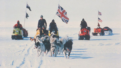 Arctic expedition with dog sled team and skidoos carrying British and American flags across snowy polar ice.
