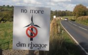 Roadside sign reading 'no more'/'dim rhagor' and a wind turbine crossed out, opposing new wind farms in a rural countryside setting.