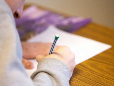 A student writes on an exam paper whilst being sat at a wooden desk.