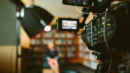 A person sitting in front of a bookcase and a camera set up for an interview.