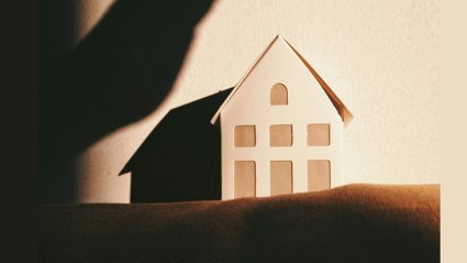 Shadow of a hand looming over a small paper house on a textured surface.