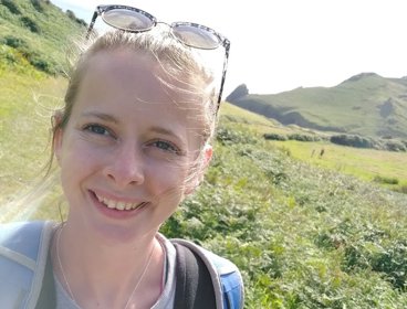 Person wearing sunglasses and a backpack, standing on a sunny grassy hillside with rolling green mountains in the background.