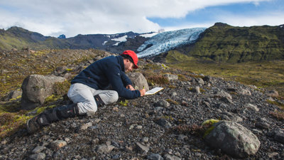 A researcher laying on the rocky ground taking notes. In the background is a mountainous landscape.
