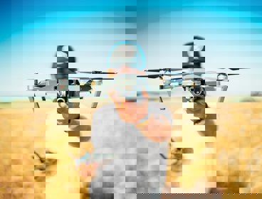 A man standing in yellow field of wheat, holding a silver drone out towards the camera. The drone obscures his face.