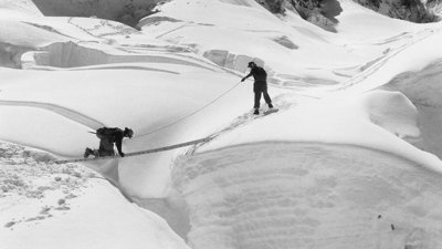 Black and white photo of two explorer in a desolate, snowy area crossing a gap using a ladder horizontally.