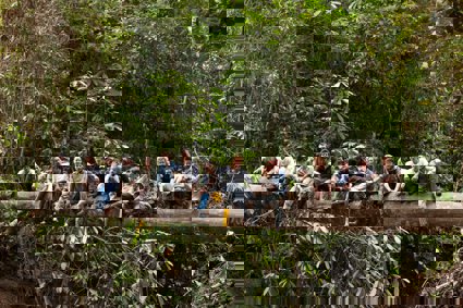 Group of people sitting on a large horizontal tree trunk surrounded by forest.
