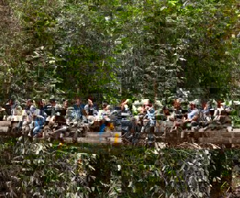 Group of people sitting on a large horizontal tree trunk surrounded by forest.
