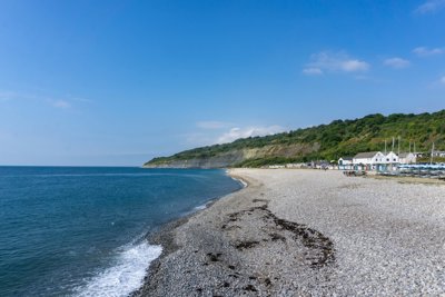 A grey pebble beach in Lyme Regis, UK, streatches accross the foreground with beach houses in the middle ground and grass covered rocky hills in the background.