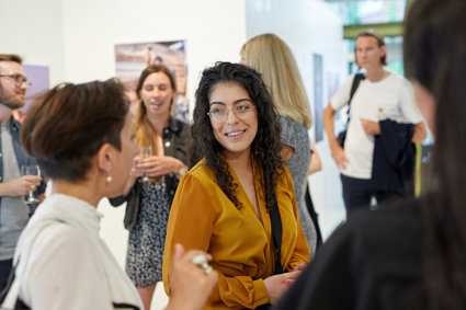 A person stands amongst a group of people chatting and drinking in a brightly lit exhibition space.