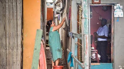 A person hangs up wet clothes on a washing line outside of the house whilst a person on the other side of the wall, inside the house stands and stares at the camera.