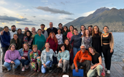 Large group of students posing for picture on pier on lake against backdrop of mountains. 