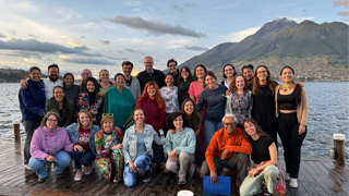 Large group of students posing for picture on pier on lake against backdrop of mountains.