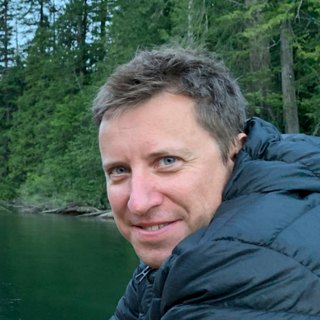Headshot of Christopher Walker in a thick, padded coat. In the background are a river and a forest.