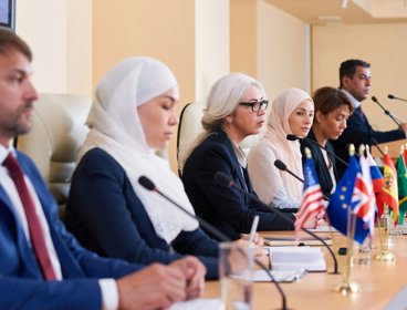 Delegates at an international conference sat at a wooden table with microphones and flags in front of them.
