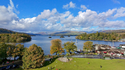 Lake Windermere surrounded by trees, distant hills, and a park area in the foreground on a sunny day.