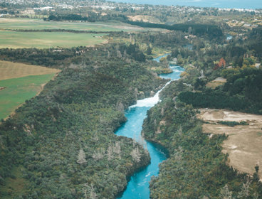 Aerial view of a winding blue river cutting through green forest and farmland with a town in the distance.