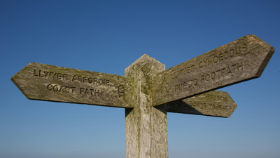 Wooden signpost showing three directions in English and Welsh. The left arrow says 'coast path' and the right arrow says 'public footpath'.