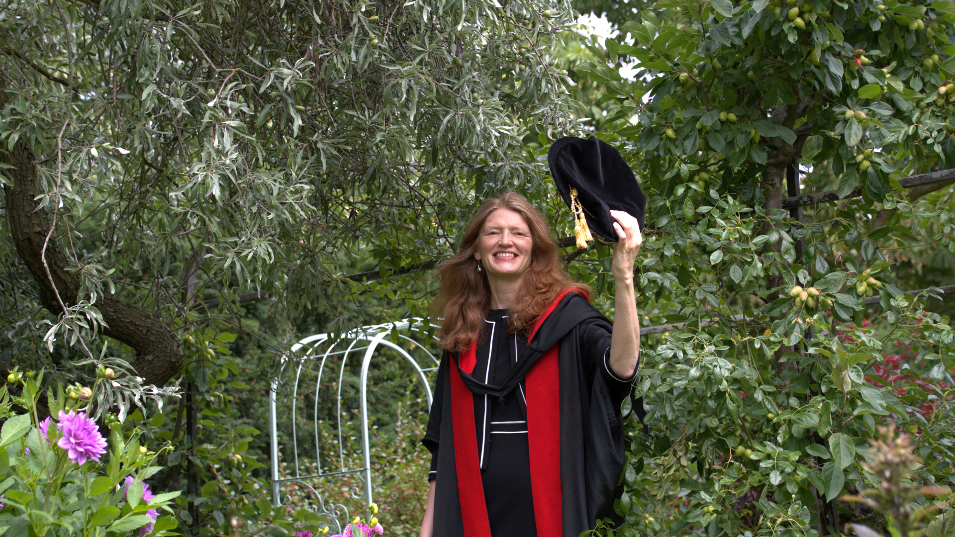 Susan Smith in graduation robes, about to throw their graduation cap. They are smiling and standing in a green, lush garden.