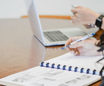 One person sits before a notebook and holds a pen whilst another sits in front of a laptop.