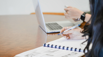 Two people sit next to each other at a wooden table. one sits before a notebook and holds a pen whilst the other sits in front of a laptop