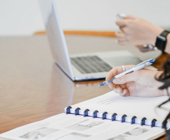 Two people sit next to each other at a wooden table. one sits before a notebook and holds a pen whilst the other sits in front of a laptop