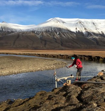 A researcher collecting water temperature measurements at the edge of a river in a baron landscape, with mountains in the background. 