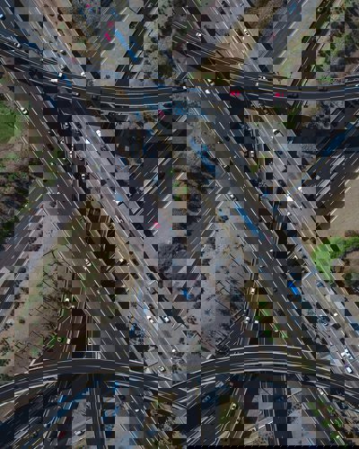 An aerial view of a motorway junction showing the roads crossing each other in an attractive pattern. Some roads are straight, others are curved. Each has multi coloured cars on it.