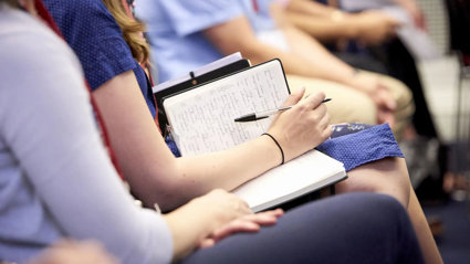 Audience member with a notebook and pen on their lap, seated next to other people.