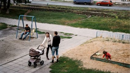 A playground with two adults with a pram and a young child playing in a sandbox.
