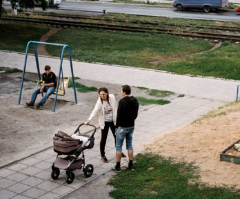 A playground with two adults with a pram and a young child playing in a sandbox.