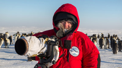 Portrait photo of Sue Flood in the arctic with winter gear and a large camera. A penguin colony stands in the background.