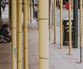 A person sitting on pavement which features scaffolding, a black bin and a bus stop.  The image suggests this person is homeless.