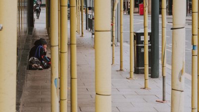 A person sitting on pavement which features scaffolding, a black bin and a bus stop.  The image suggests this person is homeless.