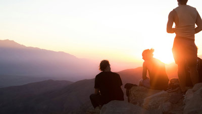 Two people sitting and one person standing near cliff taken during golden hour.