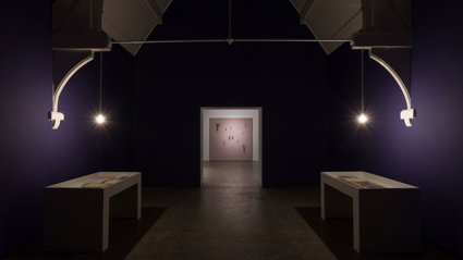 Dimly lit gallery room with deep purple walls and two white display tables.