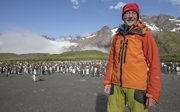 Man posing in front of a large group of pinguins and a mountain range in the background.