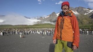 Man posing in front of a large group of pinguins and a mountain range in the background.