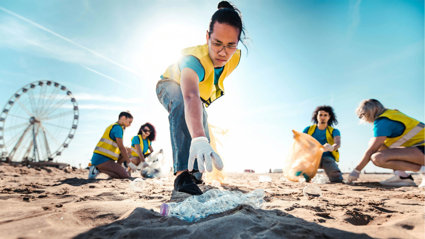People picking up litter on a beach on a sunny day. In focus is a person reaching for a plastic bottle.