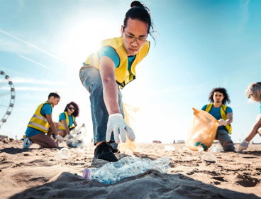 People picking up litter on a beach on a sunny day. In focus is a person reaching for a plastic bottle.