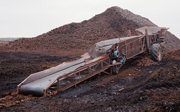 A rust-coloured conveyor machine on muddy ground beside a large pile of rectangular peat blocks.