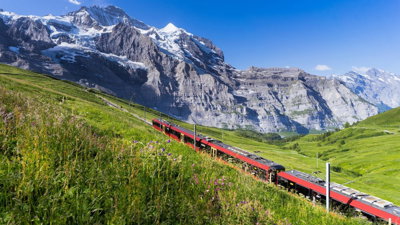 Red train in the Swiss Alps, with mountains behind and green fields in front.