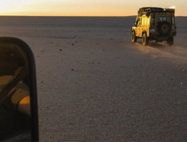 A person reflected in the car mirror, with cars in front driving across the desert.