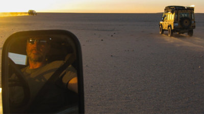 A person reflected in the car mirror, with cars in front driving across the desert.