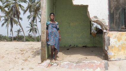 A local resident of Fuvemeh, Ghana, standing on a beach while leaning against the wall of a building damaged by coastal erosion.