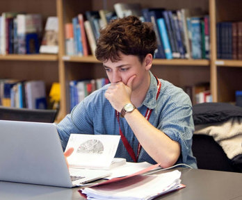 Person sat at a table looking at a laptop screen with notepads and books surrounding the laptop. Behind the table there are bookshelves with stacks of books.