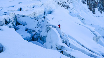 A mountain side covered in a thick layer of snow and ice, with in the distance a lone climber.