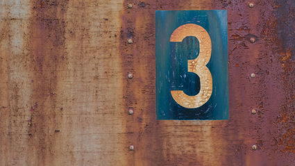 An orange coloured number 3 on a blue background, on a further rusty metal background.