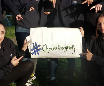 A group of students are holding and pointing at a sign that reads 'Choose Geography'.